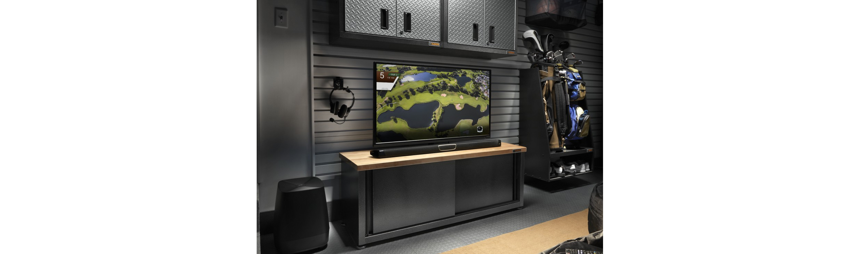 A garage with a TV on a Gladiator Cabinet below a Gladiator Wall Storage Unit. Next to the TV is a storage unit with golf clubs, backpacks and shoes. Hanging on a Gladiator Hook are headphones