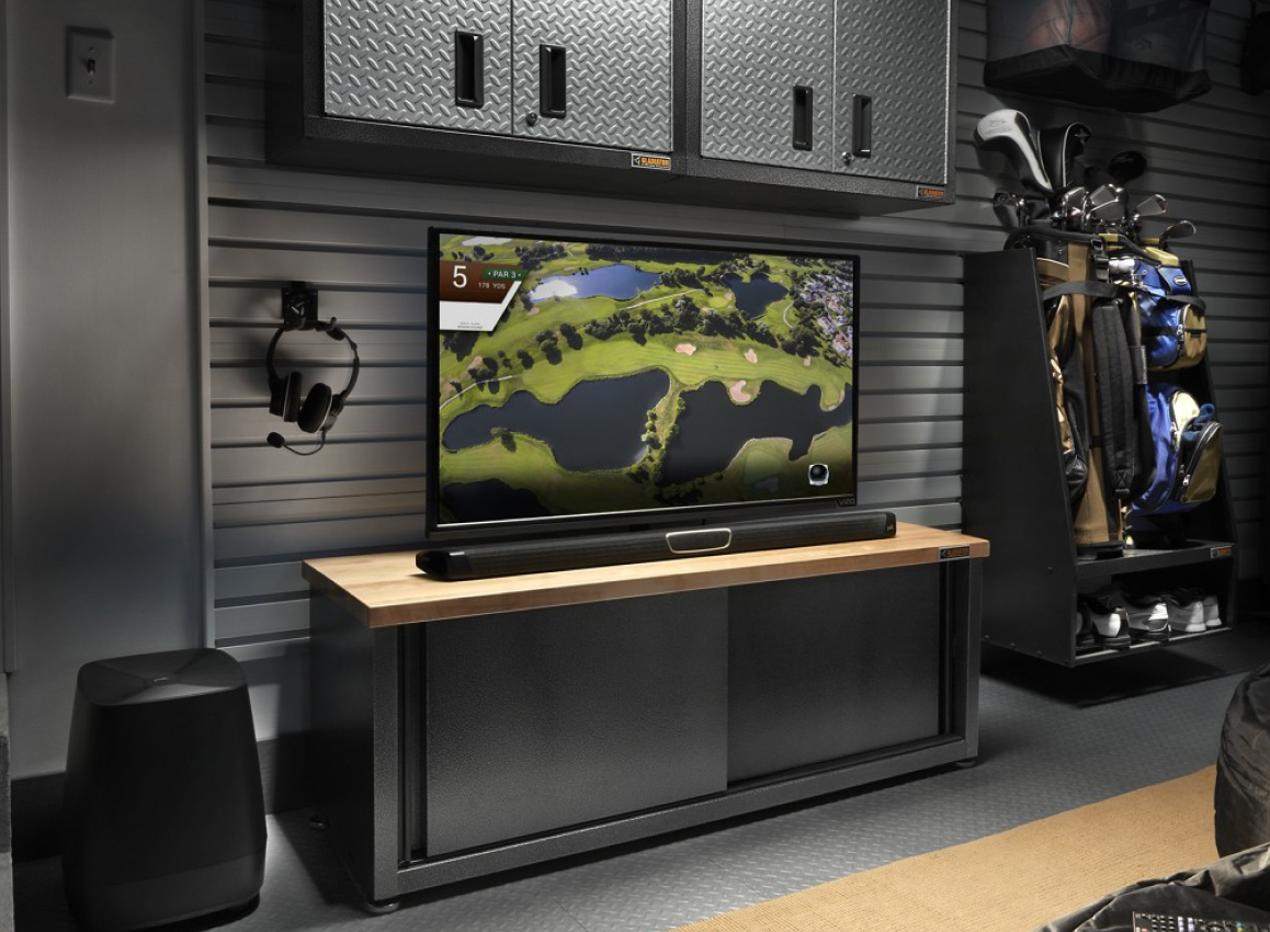 A garage with a TV on a Gladiator Cabinet below a Gladiator Wall Storage Unit. Next to the TV is a storage unit with golf clubs, backpacks and shoes. Hanging on a Gladiator Hook are headphones