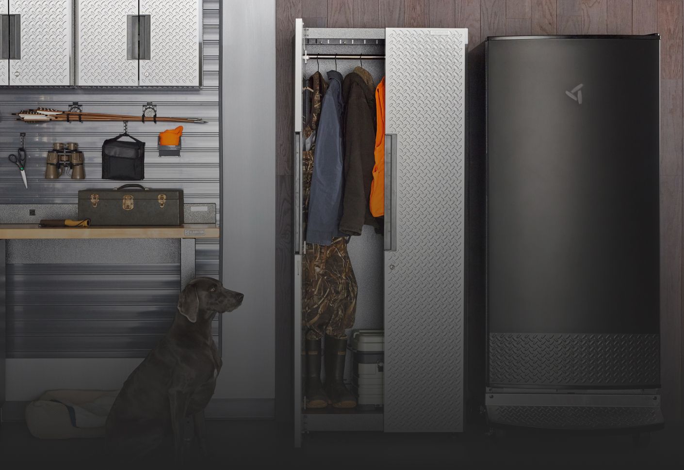 A garage with a sitting dog. A Gladiator Cabinet with one door opened. Inside are hanging jackets. Next to the cabinet is a Gladiator Garage Refrigerator